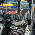 Interior of a MANITOU operator cab with steering wheel, seat, and control panel; orange warning label visible, outdoor heavy equipment lot beyond a window.