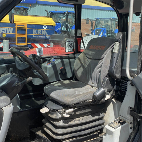 Interior of a MANITOU operator cab with steering wheel, seat, and control panel; orange warning label visible, outdoor heavy equipment lot beyond a window.