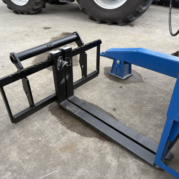 Forklift pallet fork attachment on a concrete lot, with a blue lift frame to the right and large tires in the background.