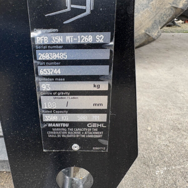 Close-up of a black machinery data plate with designation, serial and part numbers on a dusty surface.