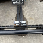 Top-down view of forklift forks resting on a trailer bed, with a Stewart sign and muddy boots at the edge.