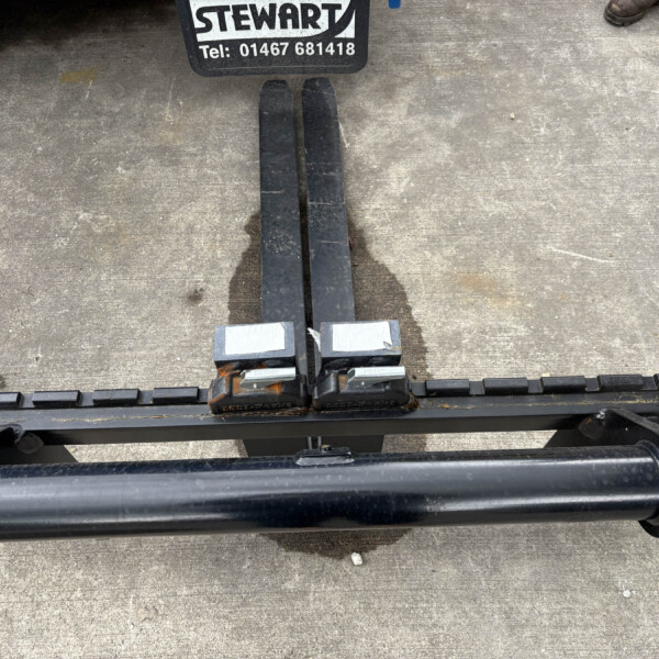 Top-down view of forklift forks resting on a trailer bed, with a Stewart sign and muddy boots at the edge.