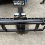 Forklift forks attached to a pallet carriage on a concrete surface, with a Stewart sign and phone number visible above; worker boots nearby.