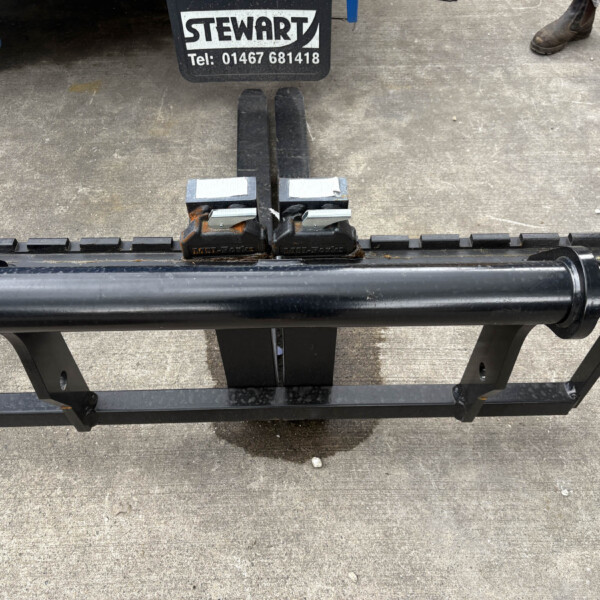 Forklift forks attached to a pallet carriage on a concrete surface, with a Stewart sign and phone number visible above; worker boots nearby.