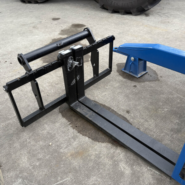 Black metal pallet fork carriage with two long forks lying on a concrete surface, part of a blue lift nearby, outdoor setting.
