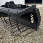 Black skid-steer grapple rake with curved tines resting on gravel outside a warehouse.