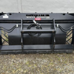 Large black bucket attachment for a skid-steer loader, with hydraulic hoses and metal arms, resting on gravel against a gray wall