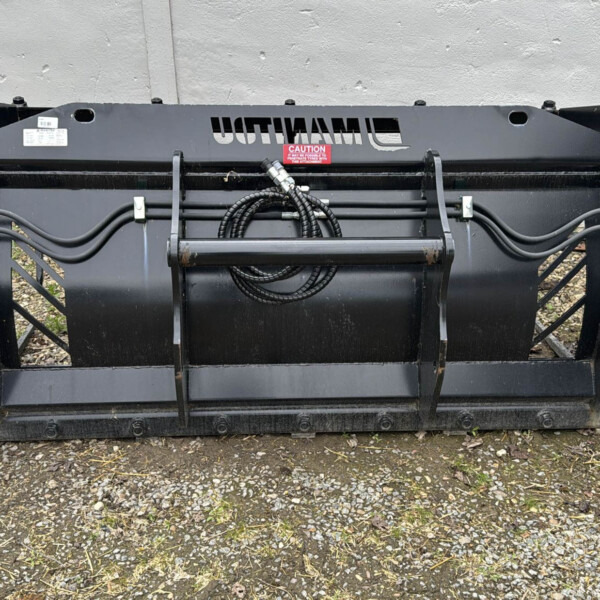 Large black bucket attachment for a skid-steer loader, with hydraulic hoses and metal arms, resting on gravel against a gray wall