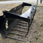 Large black skid-steer bucket attachment with teeth, resting on a gravel surface beside railroad tracks.