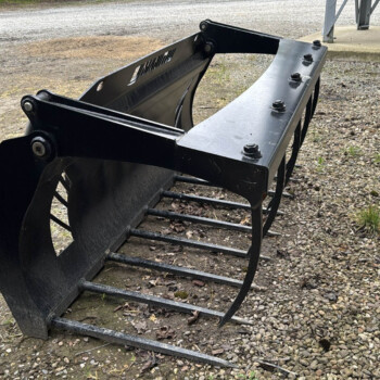 Large black skid-steer bucket attachment with teeth, resting on a gravel surface beside railroad tracks.