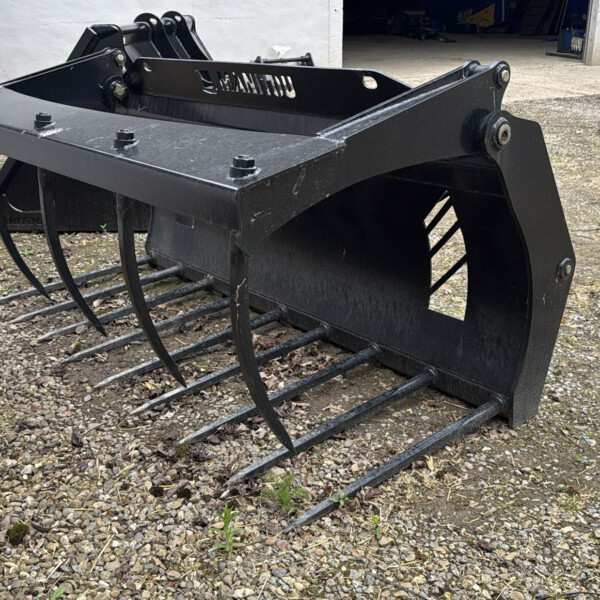 Black skid-steer grapple rake with curved tines resting on gravel outside a warehouse.