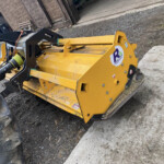 Yellow heavy machinery attachment mounted to a loader, showing hydraulic hoses and a black mounting arm on a gravel surface.