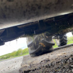 Underneath a heavy vehicle, showing a tire and the undercarriage with grass stuck to it on a gravel road.