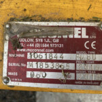 Close-up of a red/white manufacturer data plate on yellow machinery, with dirt and grass fragments obscuring the label and parts of the text visible (serial, model, Made in England).