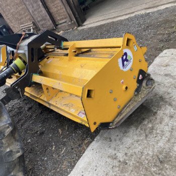 Yellow heavy machinery attachment mounted to a loader, showing hydraulic hoses and a black mounting arm on a gravel surface.