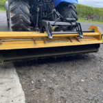 Blue tractor with a yellow front-mounted implement attached, resting on a gravel driveway with hedges in the background
