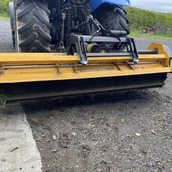 Blue tractor with a yellow front-mounted implement attached, resting on a gravel driveway with hedges in the background