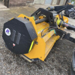 Close-up of a yellow industrial loader attachment with a black bucket and hydraulic arms on a gravel lot. In the background are a building and a parked car.