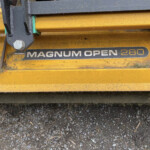 Close-up of a yellow Magnum Open 280 agricultural implement with a black mounting bracket and a large bolt, on a gravel surface.