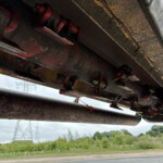 Close-up of rusted red-painted industrial machinery underside with bolts and steel beams outdoors at a construction site.
