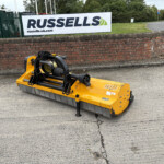 Yellow industrial road sweeper attachment parked on a road beside a brick wall and sign partly visible in the background.