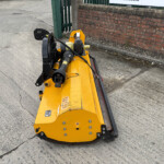Yellow construction trencher attachment on a concrete surface beside a brick wall, with visible hydraulic hoses and controls in the foreground