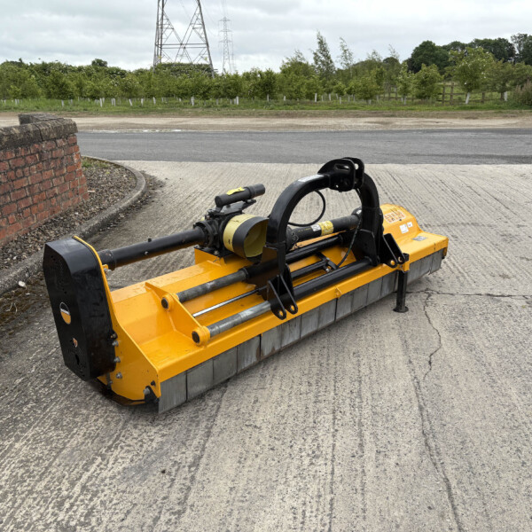 Yellow road milling machine on cracked concrete beside a brick wall, with trees and power lines in the background and a cloudy sky.