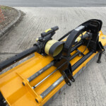 Close-up of a yellow construction attachment with black hydraulic components and a large black pipe, resting on a paved surface beside a brick wall edge.