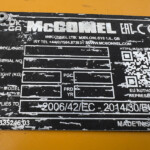 Close-up of a worn black identification plate on a yellow surface; prominent 'MCGONNEL' text, with a QR code and contact details visible.