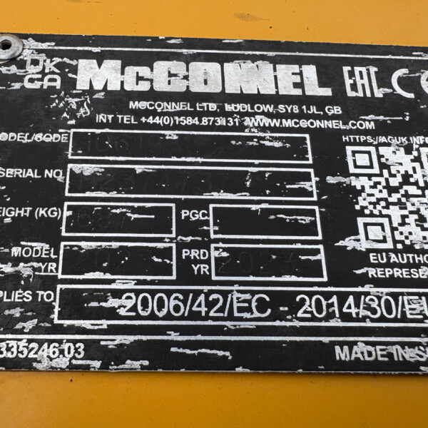 Close-up of a worn black identification plate on a yellow surface; prominent 'MCGONNEL' text, with a QR code and contact details visible.