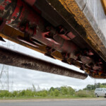 Close-up of a rusted excavator attachment with red and yellow metal, teeth and hydraulic parts, viewed from beneath against a cloudy sky and distant car
