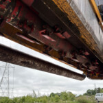Close-up of a heavy construction machine's underside showing rusted red beams and yellow supports outdoors on a cloudy day