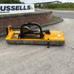 Yellow street-sweeper/brush attachment on a paved road next to a brick wall and a Russell's sign in the background.