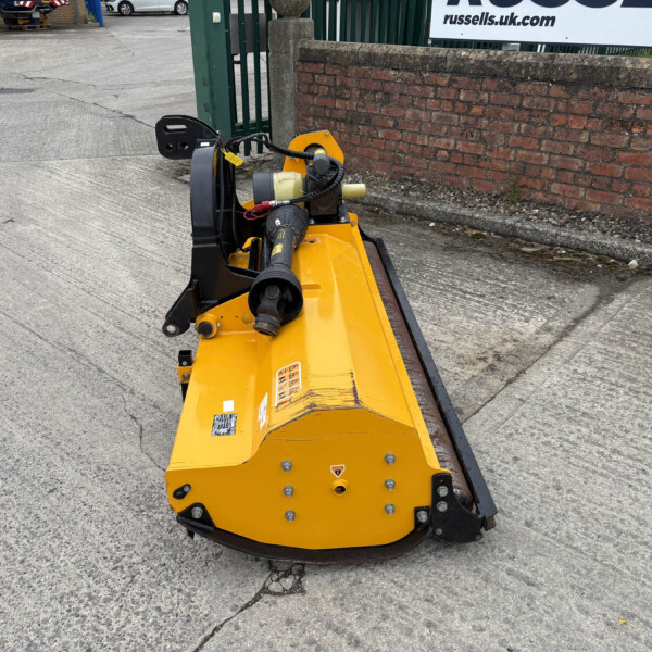 Yellow industrial compactor with hydraulic hoses parked beside a brick wall at an outdoor work site, near a green gate.