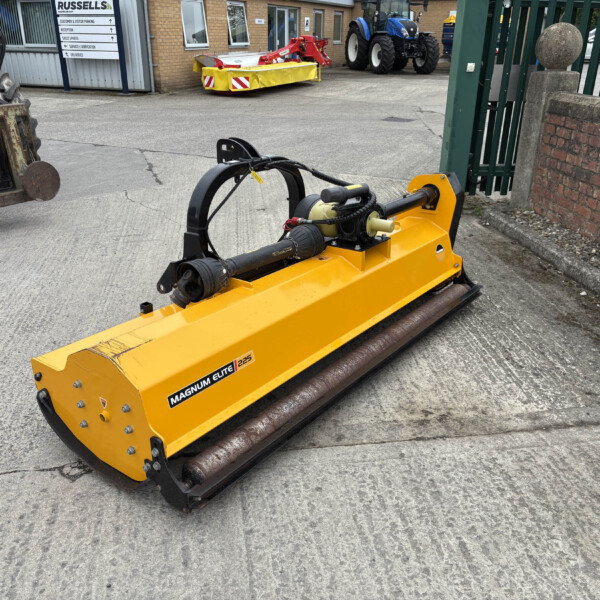 Yellow Magnum Elite 225 industrial machine with hydraulic arms, in a yard with tractors and a rusty forklift in the background.