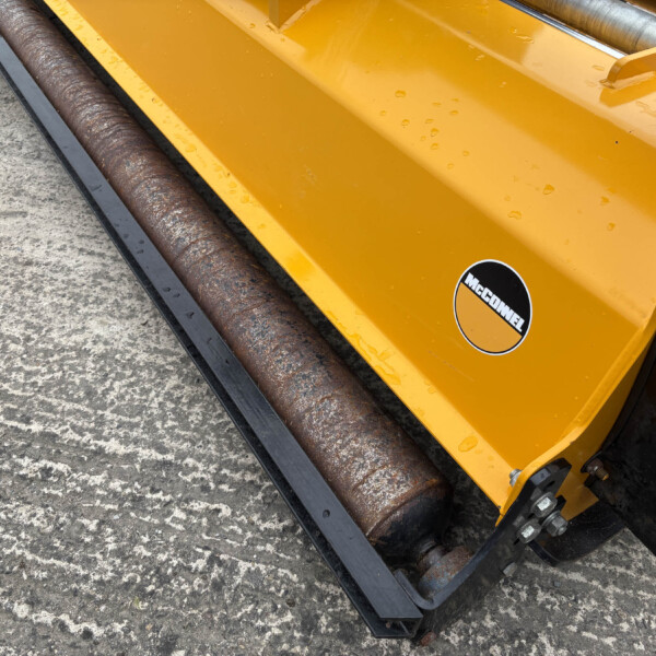 Close-up of a yellow industrial machine with a rusted cylindrical roller resting on a concrete surface, a McConnel sticker visible on the yellow frame.