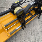 Close-up of a yellow construction attachment with black hydraulic arm and hoses, mounted on concrete surface, likely a concrete cutting tool