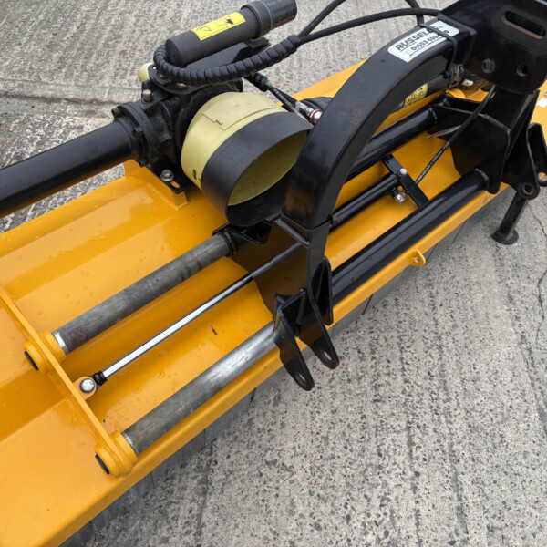 Close-up of a yellow construction attachment with black hydraulic arm and hoses, mounted on concrete surface, likely a concrete cutting tool