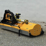 Yellow hydraulic construction attachment on a tracked machine sits on gravel beside a concrete wall, ready for use.