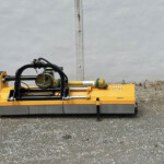 Orange hydraulic construction attachment with hoses and rotating mechanism on a gravel surface in front of a gray concrete wall