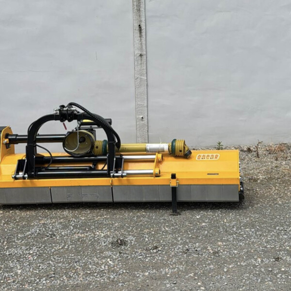 Orange hydraulic construction attachment with hoses and rotating mechanism on a gravel surface in front of a gray concrete wall