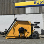 Yellow industrial machine with large blades and wheels parked on gravel in front of a corrugated metal wall with a blue-yellow sign above it.