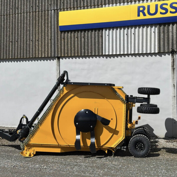 Yellow industrial machine with large blades and wheels parked on gravel in front of a corrugated metal wall with a blue-yellow sign above it.