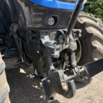 Close-up of a blue tractor's front end, showing the grille, round headlight housing, and heavy hydraulic hitch with large tires nearby.