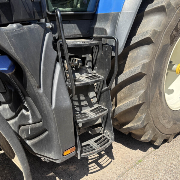 Close-up of a farm tractor's step ladder and handle near a large rear tire, with a blue fuel cap visible on the left.