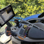 Cockpit of a heavy vehicle with a mounted tablet showing dashboards, a joystick, and numerous control panels, with a sunny windshield and leafy background outside.