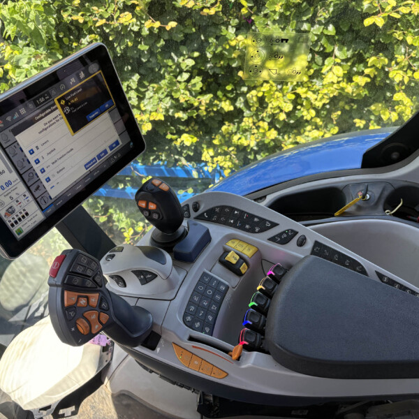 Cockpit of a heavy vehicle with a mounted tablet showing dashboards, a joystick, and numerous control panels, with a sunny windshield and leafy background outside.