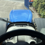 View from inside a vehicle cab looking forward at a blue hood and windshield; open blue gate ahead with hedges on the right side.