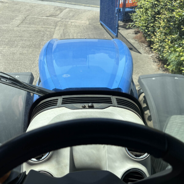 View from inside a vehicle cab looking forward at a blue hood and windshield; open blue gate ahead with hedges on the right side.
