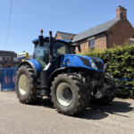 Blue New Holland tractor parked on a street in front of a brick house with a hedge and blue gate.